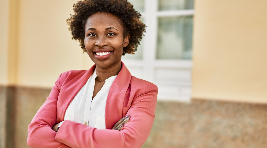 A woman in a pink suit crosses her arms and smiles.